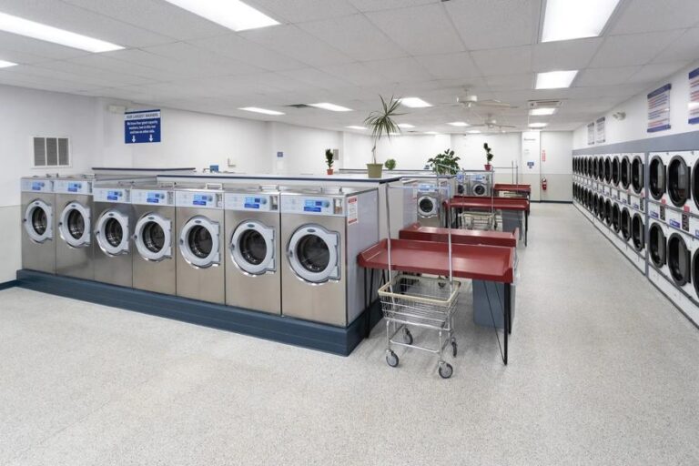 Rows of front-load washers and stacked dryers with folding tables and laundry carts in a coin laundry