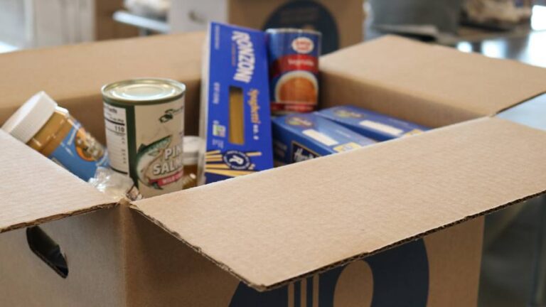 Open cardboard box of pantry groceries with pasta, canned salmon, peanut butter and soup